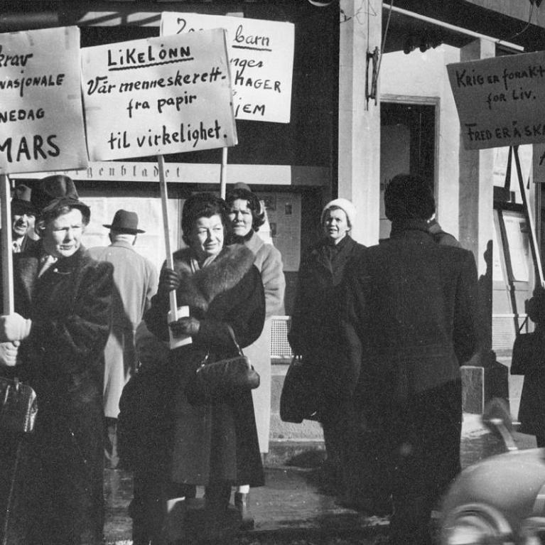 Åtte kvinner går langs fortauet med plakater på det som trolig er Norges første 8. mars markering. Parolene handler om likelønn, fred og nei til atomvåpen Åtte kvinner går langs fortauet med plakater på det som trolig er Norges første 8. mars markering. Parolene handler om likelønn, fred og nei til atomvåpen