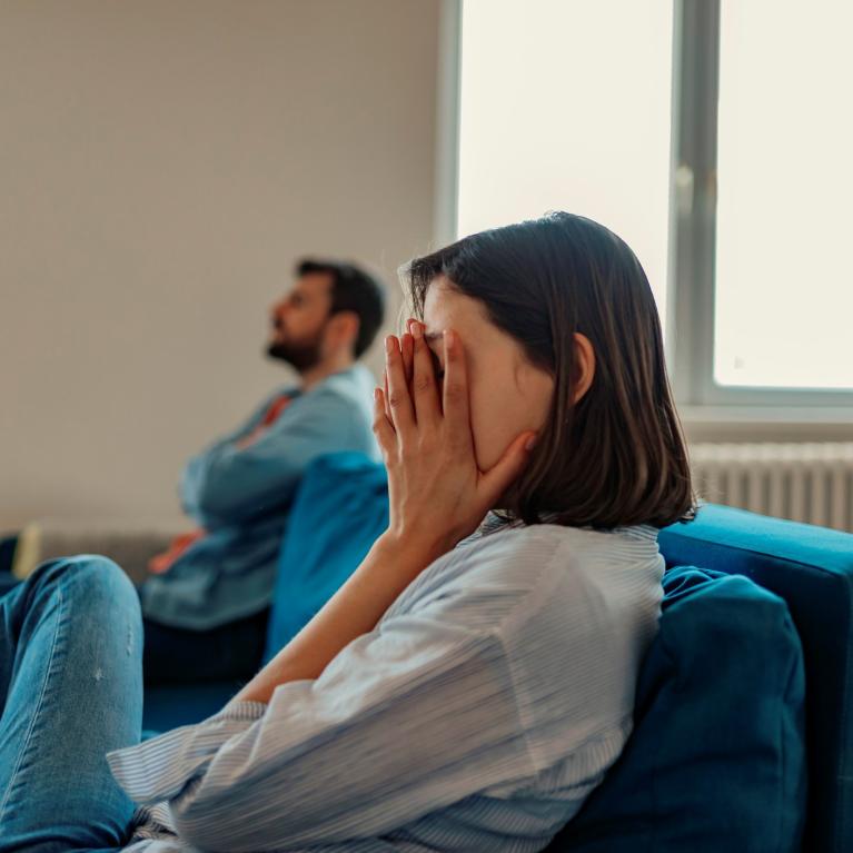 Kvinne og mann i sofa Kvinne i blå sofa med hendene foran ansiktet. Rådvill mann i bakgrunnen.
