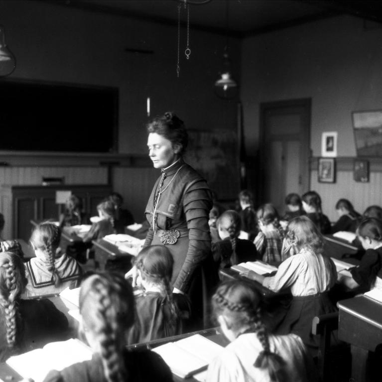 Anna Rogstad i klasserommet i 1909 Sort-hvitt-foto av Anna Rogstad stående i klasserommet mens barna sitter bøyd over pultene sine i 1909