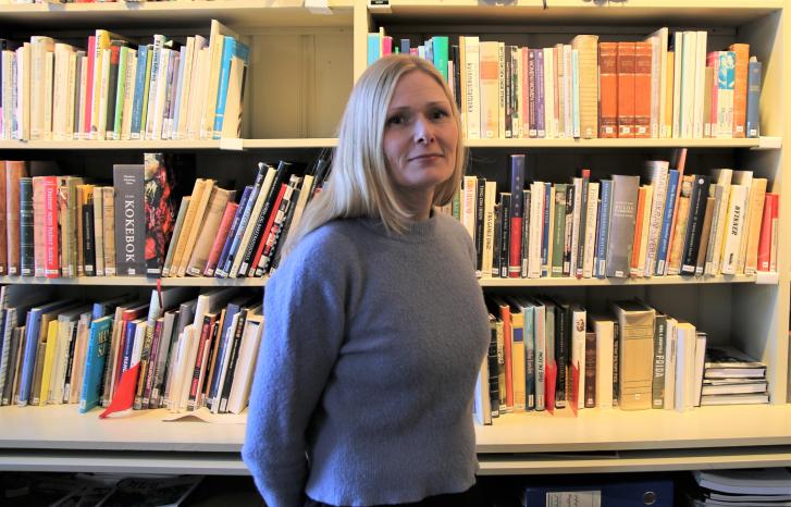 Ung kvinne med lyst, langt hår i blå genser, stående foran en bokhylle i Kvinnemuseet.