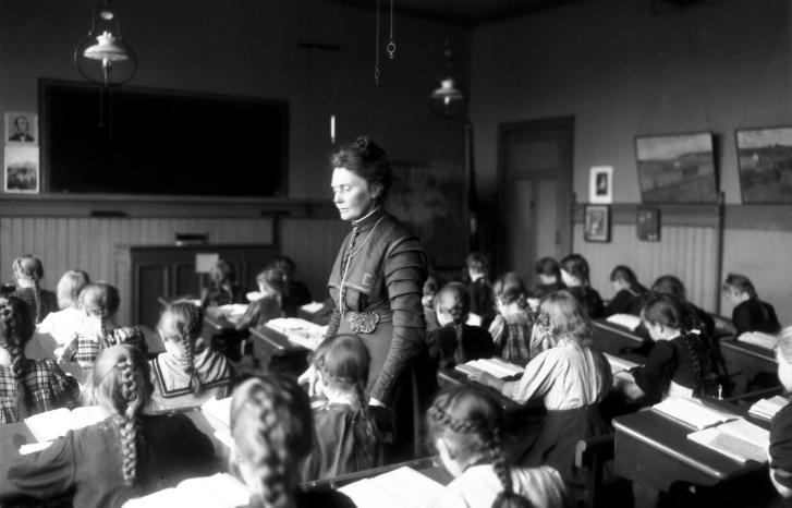 Sort-hvitt-foto av Anna Rogstad stående i klasserommet mens barna sitter bøyd over pultene sine i 1909