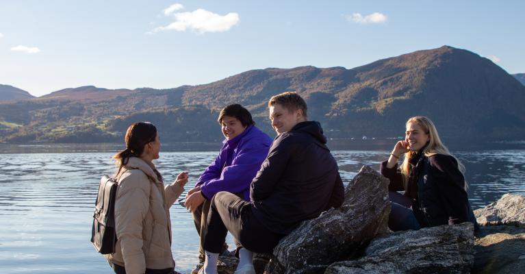 En gruppe unge mennesker sitter ved vannet med fjell i bakgrunnen