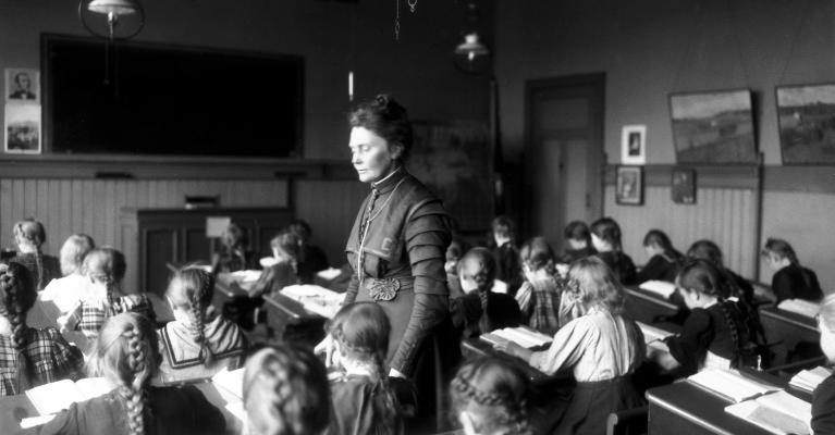 Sort-hvitt-foto av Anna Rogstad stående i klasserommet mens barna sitter bøyd over pultene sine i 1909