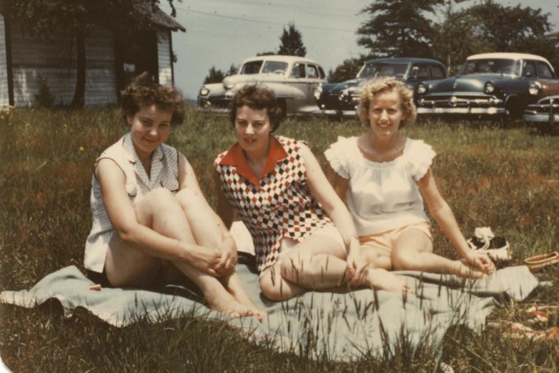 Tre kvinner fotografert i New Jersey på 1950-tallet, i sommerklær sittende på et teppe på en gresslette med amerikanske biler parkert i bakgrunnen