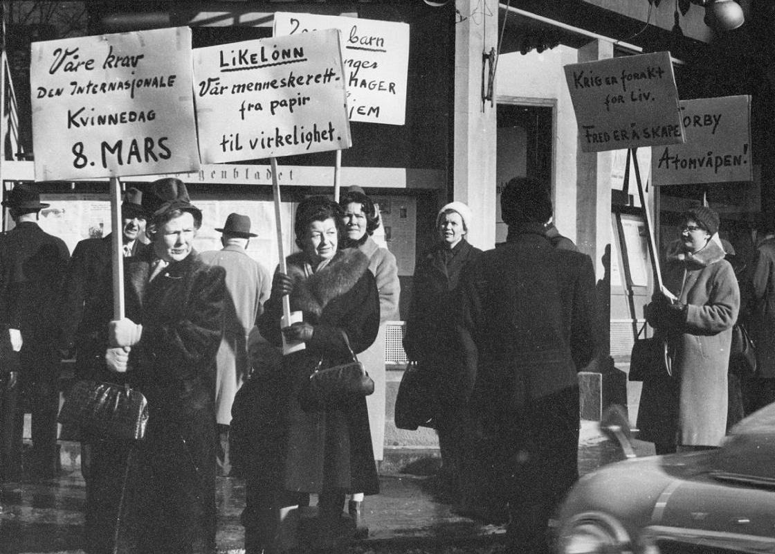 Åtte kvinner går langs fortauet med plakater på det som trolig er Norges første 8. mars markering. Parolene handler om likelønn, fred og nei til atomvåpen