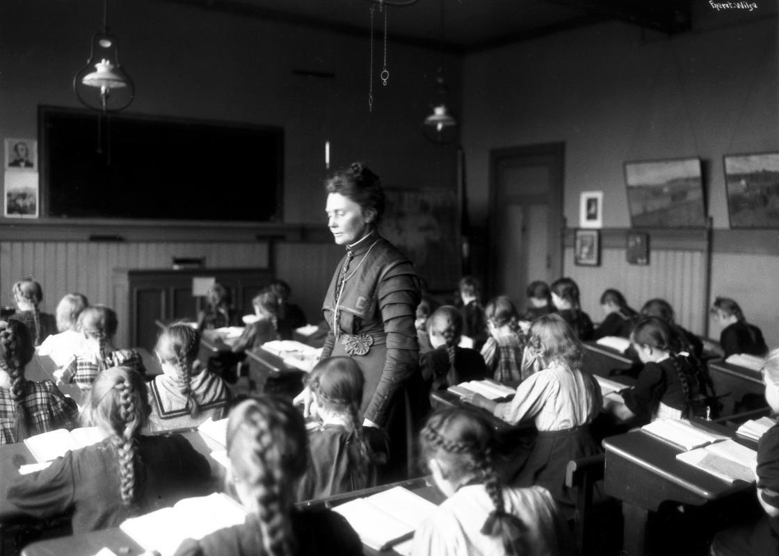 Sort-hvitt-foto av Anna Rogstad stående i klasserommet mens barna sitter bøyd over pultene sine i 1909