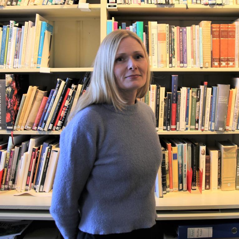 Eva Refsahl Ung kvinne med lyst, langt hår i blå genser, stående foran en bokhylle i Kvinnemuseet.