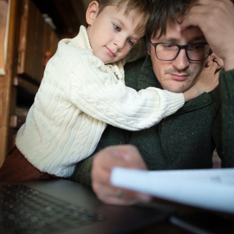 Mann sitter foran en dataskjerm og holder et ark, og ser stresset ut. Et barn holder rundt ham. Mann sitter foran en dataskjerm og holder et ark, og ser stresset ut. Et barn holder rundt ham.