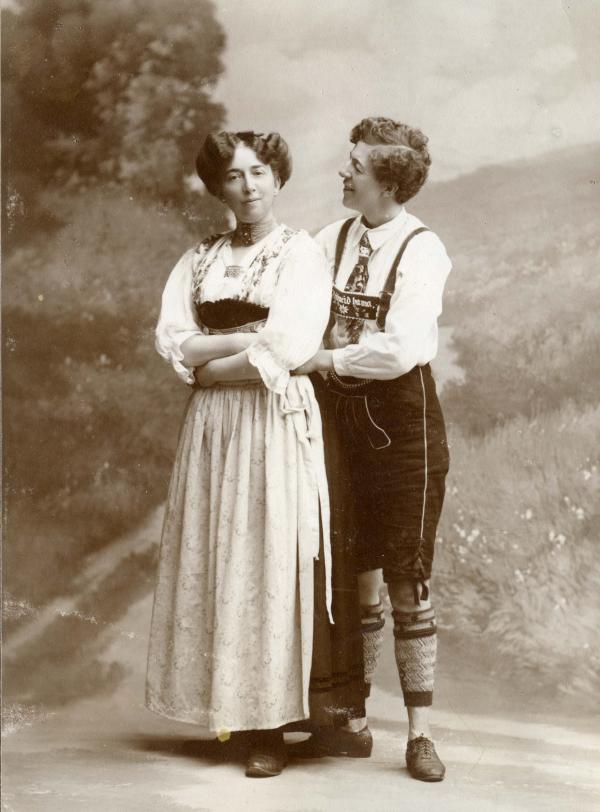 Two women holding around each other in a black and white photo, one is dressed in traditonal women's clothes, the other in men's clothes